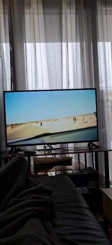 A person is lying on a couch, watching TV. The TV screen shows a scene of a car driving through a desert landscape. A person with a blue head covering is leaning out of the car window. The background includes light gray curtains. Underneath the TV is a glass table with several electronic devices.