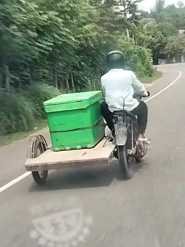A person wearing a light blue shirt and a dark helmet is riding a motorcycle with a sidecar. The sidecar is carrying a large, green box secured with straps. The motorcycle and sidecar are traveling on a paved road that appears to be in a rural or suburban area. The road has a white line marking the lane. There are trees and vegetation on the left side of the road, and some buildings and power lines are visible in the background. The video appears to be filmed from a moving vehicle behind the motorcycle. The lighting suggests it is daytime, likely overcast.