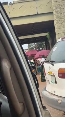 The video shows a street scene from the interior of a vehicle. In the foreground, the side panel and window frame of the vehicle are visible. Beyond the vehicle, a street market is set up under an overpass. Two individuals, likely vendors, are visible behind market stalls. One person is wearing a white shirt and appears to be interacting with a customer or arranging goods. The other individual, wearing a dark shirt, is also behind a stall. Several red market umbrellas are set up to provide shade. Merchandise is visible on the stalls. The environment appears to be a busy urban area with traffic. A white vehicle, possibly a minivan, is parked in front of the market stalls. The overpass structure is made of concrete and brick. The ground appears to be paved. The lighting suggests it is daytime.