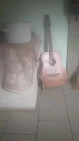 The video displays a dimly lit room with tiled walls. A light-colored acoustic guitar is propped up against a wall. In the foreground, a folded blanket or fabric is placed on a white surface. A grey cylindrical object is visible in the lower left corner of the frame. The image is tilted and somewhat blurry, making it difficult to discern fine details.
