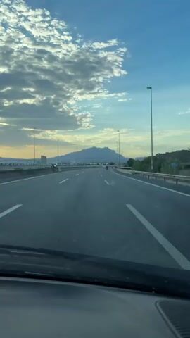 The video shows a view from the inside of a car driving on a highway. The road is paved with asphalt and has white lane markings. There are several other cars on the road, including a white SUV that is in front of the camera car. The sky is blue with scattered clouds, and there are mountains in the distance. The sun appears to be setting or rising, casting a warm light on the scene. There are streetlights along the side of the road. The dashboard of the car is visible at the bottom of the frame.