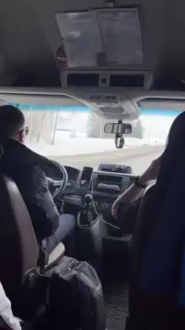 The interior of a van is shown in a video. The driver wears a black jacket and drives down a road lined with snow and trees. There is a passenger in the front seat, and a backpack on the floor. The dashboard has a phone, some controls, and documents attached to the roof. Music plays in the background.