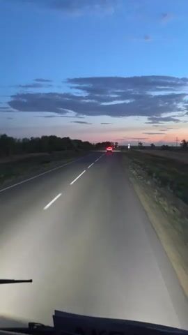 This media file captures a drive on a road in Baiqadam, Kazakhstan, during dusk. The sky is a deep blue, transitioning to hues of pink and orange near the horizon, with scattered clouds. The road, a paved two-lane highway with a white dashed line, stretches forward into the distance.

The primary objects in motion are vehicles on the road. The perspective is from inside a vehicle, likely a bus or RV, with a windshield wiper visible in the foreground. Headlights from the recording vehicle illuminate the road. A dark-colored car passes by on the left lane, its headlights also visible. Further ahead, a red car with illuminated taillights is traveling in the same direction, followed by other vehicles, their headlights visible in the distance.

The setting is a rural landscape with sparse vegetation and fields on either side of the road. Trees line the left side of the road in the mid-ground. The overall impression is one of travel as the day ends, with the soft glow of the sunset and the artificial light of vehicle headlights creating a serene yet active atmosphere.

No people are clearly visible. The activity is driving on a highway at twilight. There are no discernible emotions conveyed, beyond the inherent peacefulness of a sunset drive.

Visible text includes "IKEA" on the dashboard of the recording vehicle. There are also road signs visible in the distance, though the text on them is not legible.