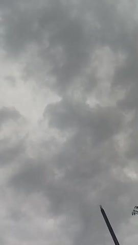 The media file captures a cloudy sky from a low-angle perspective, likely in Mubi, Nigeria. The dominant subject is the sky filled with dense, grey clouds, suggesting overcast weather, possibly before or during rain.

In the lower right corner, a dark, slender object extends upwards into the frame. It appears to be a pole or mast, possibly part of a street lamp or a tall structure. A small, bright white light source is visible at its apex, indicating it is a man-made object. The overall scene evokes a somber or dramatic mood due to the heavy cloud cover. The time of day is not definitively ascertainable but the light suggests it is daytime, though the clouds obscure direct sunlight. There is no visible text in the media file. The setting is likely an outdoor urban or semi-urban environment in Mubi, Nigeria, given the presence of what appears to be man-made infrastructure against a natural sky. No people are visible, and the activity taking place is simply the passive observation of the sky and its atmospheric conditions.