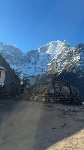 The video showcases a serene and awe-inspiring landscape in Tengboche, Nepal, featuring majestic mountains and Buddhist architectural elements under a clear blue sky.

The opening shot reveals a person in a bright neon green jacket walking along a dirt path, with a formidable snow-capped mountain range dominating the background. A stone wall with a wooden structure and a cellular tower are also visible.

The camera then pans to a small, traditional white building with a yellow curtain and an ornate golden top, situated behind a stone wall. Snow-capped peaks continue to loom in the distance.

Following this, a close-up focuses on a large, prominent white Buddhist stupa, topped with a striking golden and red striped pinnacle. A yellow fence guards its base.

The video then transitions to a panoramic view of multiple buildings with distinctive red roofs, likely part of a monastery, nestled in the mountainous terrain. A long stone wall adorned with numerous prayer wheels forms the foreground.

The final shots provide a closer look at these monastery buildings and the prayer wheel wall, set against the backdrop of a large, dark, rocky mountain. The overall impression is one of bright, sunny daylight, highlighting the rugged beauty of the Himalayas and the spiritual essence of the region. No explicit English text is clearly visible.