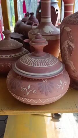 The video shows a collection of clay pottery displayed on a yellow shelf. The pottery is mostly reddish-brown with white decorative markings. In the foreground, there is a round lidded pot with intricate floral and wave-like patterns. Behind it, to the left, is another lidded pot with similar geometric designs. To the right of the central pot is a tall vase with a narrow neck and a wide base, decorated with white motifs and what appears to be a depiction of a goat or ram. Several other pottery items, including vases and bowls, are visible in the background, some of which are partially obscured. The lighting suggests an outdoor or well-lit indoor environment, possibly a market stall or artisan's workshop. The camera pans slowly across the collection, focusing on the details of the pottery.