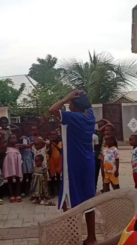 The video shows a group of children gathered in an outdoor space, likely a courtyard or patio. They are standing on a paved area with a geometric pattern. A woman in a blue and white dress is leading them in what appears to be a game or activity, with many children mirroring her actions of placing their hands on their heads. The children range in age from toddlers to older children. Behind the group, there is a building with a light-colored exterior and a metal gate with a decorative design. A tall palm tree and other greenery are visible in the background. In another segment, a woman is carrying a small child in her arms while walking past the group. A table laden with snacks and drinks is set up nearby, with some children seated around it. A few adults are also present, observing the scene. The overall atmosphere suggests a casual, social gathering, possibly a children's party or playdate.
