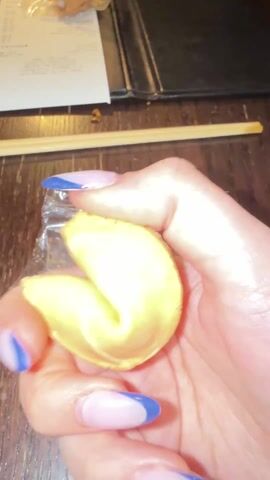 A hand with light skin and blue-tipped fingernails breaks open a fortune cookie. The fortune reads "An important lesson learned this week...".  A receipt, chopsticks and what appears to be a menu lie nearby on a dark brown wooden surface. The person has a ring with a purple stone on their finger.