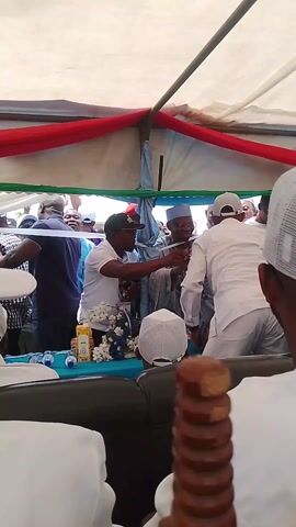 The video shows a scene under a tent adorned with red, white, and green decorations. Several people, many wearing white caps or head coverings, are visible. A man in a black baseball cap and white shirt is interacting with another man in a white shirt and cap across a table covered with a blue cloth. The table also holds water bottles, a box of juice, and a floral arrangement. There are other individuals behind them, some appearing to be engaged in conversation or observing the interaction at the table. A decorative, turned-wood object is visible in the lower right corner of the frame.