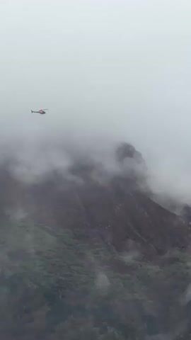 A red and white helicopter flies across a cloudy, overcast sky, moving through thick fog and clouds that partially obscure rugged, dark mountains. The helicopter is positioned relatively high in the frame, above visible mountain peaks but still amidst the swirling cloud cover. The mountains below are steep and rocky, with some patches of green vegetation visible on their lower slopes. The weather is distinctly cloudy and foggy, creating a dramatic and somewhat mysterious atmosphere. The scene depicts a remote, high-altitude mountainous region, consistent with the provided context of Namche Bazaar, Nepal. No text is visible in the video.