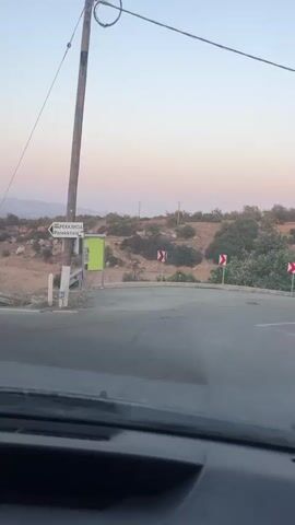 The video begins with a close-up of a road sign pointing to the right and labeled PEKKATIA and Parekklesia. The sign is attached to a wooden pole. To the right of the pole, there is a bright green rectangular sign, and further to the right, there are two curved road warning signs with red and white chevrons. The road ahead curves to the right and appears to be descending into a valley. The landscape is dry and hilly, with sparse vegetation. In the distance, mountains are visible against a pale pink and blue sky, suggesting either sunrise or sunset. The perspective suggests the video is being filmed from inside a vehicle, with the dashboard visible in the foreground. The camera pans slightly to the right, following the road as it curves. The vehicle is moving and the scene is relatively still.