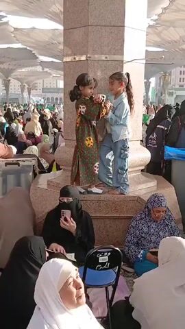 The video shows a large crowd of people, mostly women, gathered around a large pillar. Two young girls are standing on the base of the pillar, playfully interacting with each other. One girl is wearing a green and brown dress, while the other is wearing a denim jacket and pants. A young boy in a grey outfit stands close to the pillar, looking towards the girls. A woman in a niqab is sitting on a chair and holding a mobile phone, appearing to be taking a picture. Other women, wearing headscarves, are sitting around the pillar, some with books or personal items. The area is covered by a large, white, umbrella-like structure, suggesting an outdoor gathering space.
