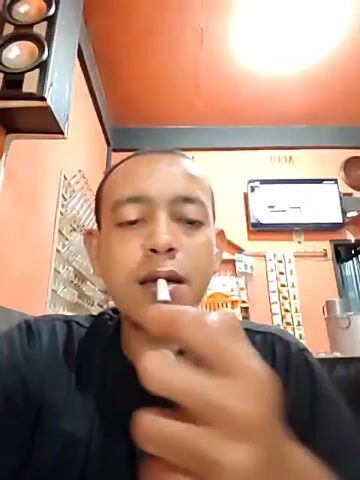 A medium shot depicts a man exhaling a large cloud of vapor from an electronic cigarette or vape device. The man is wearing a black t-shirt and is positioned indoors. Visible in the background are shelves with numerous small items, possibly glasses or decorative objects. A flat-screen television is mounted on the wall, and a set of speakers is also visible. The man’s face is clearly visible, and he looks directly at the camera after exhaling the vapor. The room has orange-colored walls, and an overhead light is illuminating the scene.