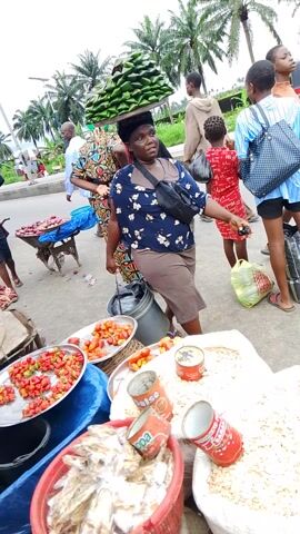A group of people are gathered at an outdoor market. In the foreground, a woman wearing a dark blue dress with a floral pattern is looking at her phone. To her right, a large white sack filled with a light-colored granular substance, possibly rice or grains, is visible. Several cans of food, some red with white labeling, are also on the ground. A young boy in a blue and orange shirt stands to the left of the woman, looking away from the camera. Behind them, another child, possibly a girl, wears a red dress. Further in the background, a market stall with various goods is set up under a blue umbrella. Palm trees line the edge of the market area, and a small vehicle, possibly a tricycle, is parked behind the stall. The ground appears to be paved. The overall impression is a bustling marketplace scene.