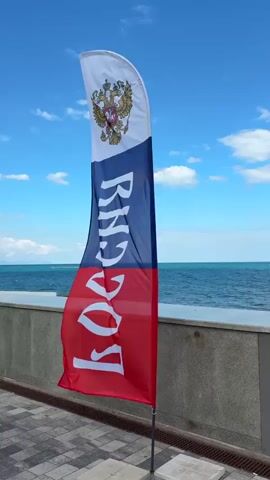 This is a close-up, eye-level, static shot of a Russian flag unfurled against a bright blue sky with scattered white clouds. The flag is positioned in the center of the frame, with a blurred expanse of the sea and a low wall serving as the background. The flag is vertically divided into three horizontal stripes: white at the top, blue in the middle, and red at the bottom. The top white section features the Russian coat of arms, a double-headed eagle with crowns. The blue and red sections bear the word "РОССИЯ" (Russia) in large white Cyrillic letters. The flag is attached to a thin, dark pole that extends downwards, with its base planted in a light-colored square concrete block, which is partially visible in the lower portion of the frame. The ground in front of the flag is paved with interlocking gray and black square tiles. The sea is calm, with gentle waves and a hazy horizon. No people are visible in the immediate vicinity of the flag. The overall impression is one of patriotism and a seaside setting.