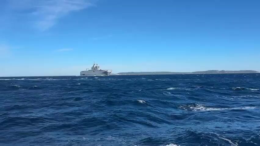 A gray military ship is sailing on a blue sea. The ship is large and has a flat deck at the front. There are buildings and hills in the background. The sky is clear and blue. The sea is choppy with waves.