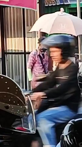 A person wearing a dark helmet and a blue hoodie is blurred as they move on a motorcycle. Behind them, a person in a light pink shirt with a patterned design and brown pants stands near a cart under a pale pink umbrella. The cart has green signage with white text that includes the word "CIK" visible. Another person wearing a mask and a cap stands behind the cart. Several motorcycles are parked around the area. The environment appears to be an outdoor market or street scene with some buildings and stalls in the background.