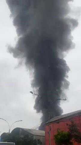 This media file depicts a large plume of thick, dark smoke rising into a cloudy sky. The smoke is so dense that it obscures the sky behind it, creating a dramatic and ominous visual. In the foreground, there are glimpses of buildings and streetlights, suggesting an urban or industrial setting. One building is partially visible on the right, with a red facade. On the left, another building with a curved roof is visible, along with trees and a streetlight. Several birds are perched on the streetlights. The overall scene evokes a sense of alarm or disaster, likely due to a fire or industrial accident. The time of day appears to be overcast, possibly late morning or afternoon, given the diffused natural light. No people are clearly visible in the frame. The location is identified as Lima, Peru. There is no visible text in the media file.
