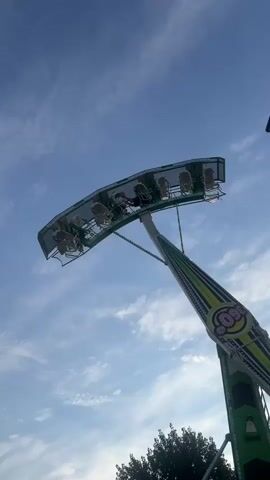 The video shows a large amusement park ride with a long arm that swings riders back and forth. The ride is green and white with the words "360 TURBO" written on it. There are several people on the ride, all appearing to be in good spirits. The ride is operating under a cloudy blue sky, with trees visible in the background. There are signs on the ride that appear to be safety instructions or pricing information. The camera is positioned at a low angle, looking up at the ride. The ride appears to be in motion, with the arm swinging. The video is short and does not contain any dialogue or sound effects.