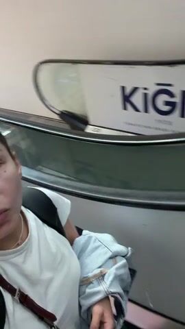 The video begins with a close-up selfie of a woman with her hair pulled back and wearing a white t-shirt and a backpack. She is on an escalator, and a logo that reads "KİĞILI 1938" is visible behind her. As the escalator moves, the scene pans out to reveal the interior of a large, modern building, likely a shopping mall or airport. The woman is in the foreground, and the environment behind her shows a multi-level structure with shops and people moving around. Another woman in a black top and jeans is visible on an adjacent escalator in the background. The video appears to be a personal recording, possibly a vlog or a travel update, showing the woman's movement through the location.