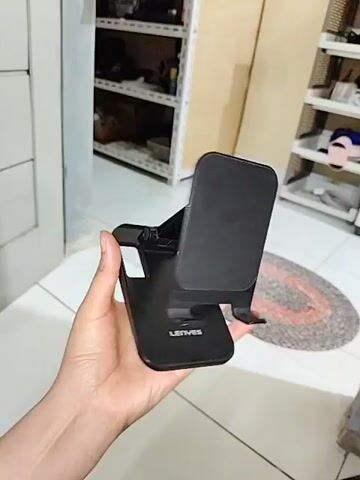 The video shows a black, foldable phone stand being held by a person's hand. The stand has a base, an adjustable arm, and a holder for a phone. The background appears to be a room with shelves and a rug. The stand is manipulated and folded in various ways to demonstrate its adjustability and portability.