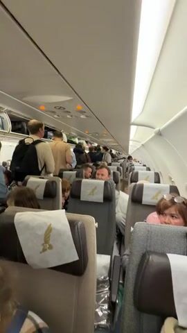 The video shows a fully occupied commercial airplane cabin with passengers standing in the aisle, apparently waiting to disembark. People are seen standing with their bags, moving slowly toward the front of the plane, while others are seated in their rows. The overhead bins appear full. The plane's interior is well-lit, with a clean, modern design, including headrest covers displaying a golden eagle emblem. The atmosphere appears organized but somewhat crowded as passengers prepare to leave the aircraft.