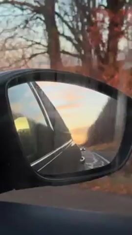 The view from a car's side mirror reflects the road behind, with another car following. The sky is a pastel mix of blues, pinks, and yellows, likely during sunrise or sunset. Trees line the roadside, and some appear bare of leaves. The mirror itself is slightly dirty, with what looks like water spots. The car is moving, as indicated by the changing scenery in the mirror.