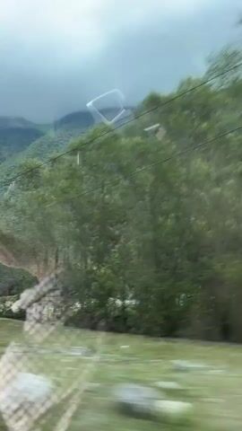 The media file captures a scenic view from a moving vehicle, likely a train or bus, through a window. The primary subject is a lush, green mountainside covered in dense forest, with a winding road visible near the summit. A bright turquoise river flows through the valley below, bordered by grassy areas. In the foreground, reflections on the window show abstract patterns, possibly from the vehicle's interior.

The overall scene depicts a journey through a natural landscape, emphasizing the grandeur of the mountains and the serenity of the river. The weather appears overcast, with a heavy, cloudy sky that casts a diffused light on the scene, suggesting it could be late afternoon or a generally gloomy day. The moving perspective creates a sense of travel and motion, with trees and foliage appearing blurred.

Notable details include what seems to be a ski lift structure on the mountainside in the latter part of the video, indicating the potential for recreational activities in the area. The presence of a fence or barrier along the riverbank suggests a managed or accessible area. The location is identified as Bzypta, Abkhazia, which provides geographical context for the mountainous terrain and river. No people are clearly visible, and no text is discernible within the media file itself. The emotion evoked is one of peaceful exploration and appreciation of natural beauty.