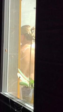 The video shows a person taking a shower through a window. The person is facing away from the camera, with their back to the viewer. They have blonde hair tied up in a bun and are holding a yellow sponge. The shower appears to be enclosed by glass and tiles. A potted plant is visible on the windowsill in front of the shower. The lighting suggests it may be evening or night.