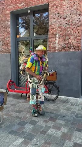 A man dressed in a colorful, patchwork outfit, including a straw hat adorned with flowers, plays a saxophone. He is standing on a paved sidewalk in front of a brick building with a large window. A red bicycle with a wooden crate attached to the back is parked behind him. The crate contains bottles. The man's attire is elaborate and includes patched pants, suspenders, and stuffed animals around his neck. His shoes are also unusual. To the right of the man, attached to the back of his bicycle, is a collection of musical instruments, including a drum and a xylophone. The man is looking forward as he plays the saxophone.