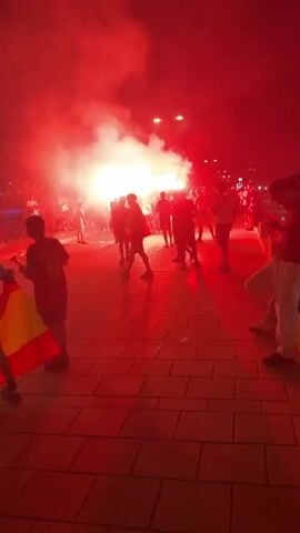 A group of people are standing on a paved walkway at night, illuminated by the bright red glow of multiple flares. Smoke from the flares fills the air, obscuring the background and casting long shadows. Some individuals are holding up phones, possibly to record the event. The people appear to be a mix of adults and possibly some younger individuals. The environment seems to be outdoors, with streetlights visible in the distance. The overall atmosphere is energetic and celebratory, with the red lighting creating a dramatic effect.