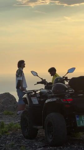 Two young men are standing next to two quad bikes on a rocky, elevated terrain overlooking the ocean at sunset. The man on the left is wearing a patterned short-sleeved shirt, blue shorts, and white sneakers. He is looking up at the sky with his head tilted back and his arms relaxed by his sides. The man on the right is wearing a yellow t-shirt and is seated on one of the quad bikes, appearing to be talking to the other man. One quad bike is black, and another is partially visible behind it. Two helmets are placed on the back of the quad bike in the foreground. The sky is a warm, hazy orange and yellow, indicating the sun is setting over the calm ocean. The ground is covered in gravel and sparse vegetation.