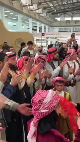 Here is a description of the video:

A group of people, mostly men, are engaged in a traditional dance, possibly Dabke, inside what appears to be an exhibition hall or large indoor event space. The men are dressed in traditional Saudi attire, including thobes, gutras (headscarves), and bishts (outer cloaks) in various colors, predominantly white, brown, and red-and-white checked patterns. Some boys and women are also participating in the dance. They are clapping their hands, moving in a coordinated manner, and appear to be enjoying themselves. A boy wears a brown thobe, and the women wear black hijabs and gowns.

The surroundings suggest a commercial setting. There are exhibition stands and booths in the background, indicating the location is likely a trade show or exhibition. People are standing around, watching and recording the performance with their phones.

A large speaker is visible, suggesting that music is playing to accompany the dance. There are decorative elements and branding visible on the stands, though the specifics are not readily discernible.