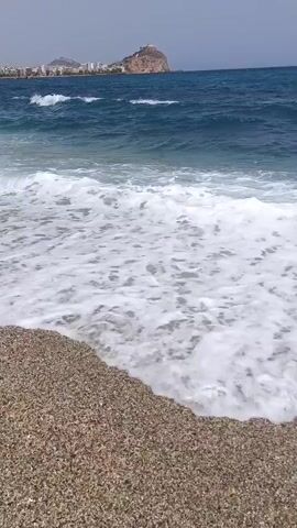 The video shows a gently sloping pebble beach with waves rolling in. The sea is a deep blue, and the sky is a clear, pale blue with a few wispy clouds. In the distance, a coastline is visible with buildings and a prominent rocky outcrop. There are no people clearly visible in the foreground, but there may be figures in the distance along the coastline. The focus is on the natural environment of the beach and sea.