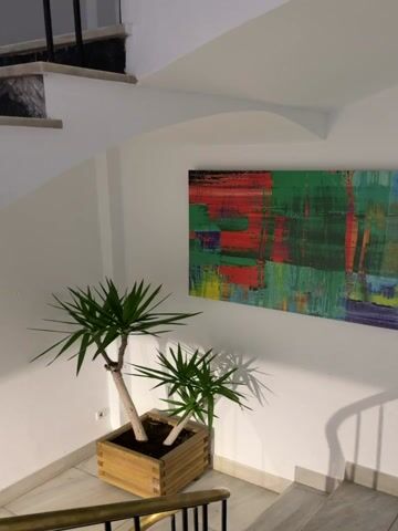 The video shows a still image of an indoor scene with a staircase. To the left, there are two potted plants with spiky green leaves, one larger than the other. The plants are in a wooden planter box. A piece of abstract art with vibrant colors, predominantly green, red, and blue, hangs on the white wall above the plants. To the right of the plants and art, a staircase with marble-like steps and a metal railing is visible. The staircase curves upwards and to the left. Shadows of the railing are cast on the white wall. The lighting suggests the scene is illuminated by artificial light.