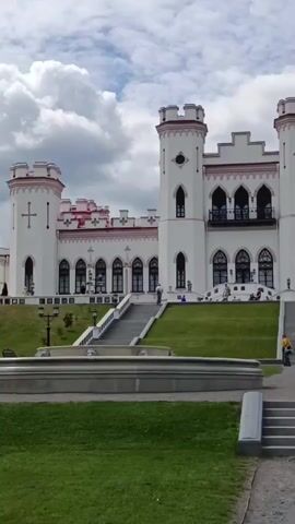 The video opens with a wide shot of a white, Gothic-style castle with red accents, featuring two prominent towers. Steps lead up to the castle entrance, and there are people visible on the steps and on the castle grounds. A fountain is visible in the foreground. The camera then pans to the right, revealing a manicured lawn, benches, and lampposts. The castle remains in the background. The pan continues, showing more of the green lawn, cypress trees, and distant trees. The camera then focuses on a statue of a woman in the middle of a garden. More cypress trees and the distant forest are visible. The video ends on a close-up of a lamppost. The sky is cloudy.