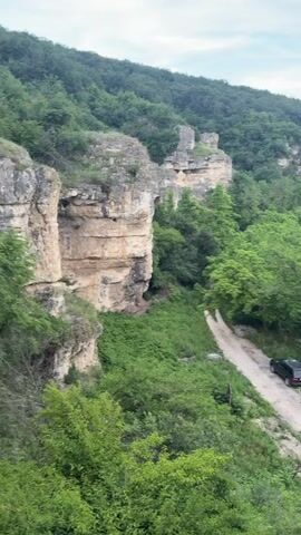 The video opens with a wide shot of a rocky canyon with a dirt road winding through it. Lush green trees and vegetation cover the canyon walls, and several large rock formations are visible. A dark-colored SUV is parked on the side of the dirt road. The camera then zooms in and pans to the right, focusing on a person climbing up a rocky incline. This person is wearing a puffer vest, dark pants, and sneakers. Another person's hand, holding a black object, is visible in the foreground, seemingly filming the climber. The background remains the rocky canyon, now out of focus.