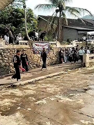 Three people dressed in dark clothing, two with patterned sashes and the third with a dark shirt, are performing a dance or martial arts routine in a wet, muddy street.  A crowd of people stands behind a low stone wall, watching.  Some are standing on the wall. A banner with text and a pattern is affixed to the wall.  Buildings and trees surround the performance space. One building is on the left, and another is partially visible on the right.  The sky is overcast.  One of the performers falls to the ground while the other two continue to move.  The street is covered in water and mud.