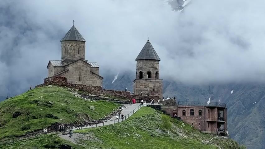 The video captures a panoramic view of Gergeti Trinity Church, a historic Orthodox church perched atop a lush green mountain. The church is made of stone and features a tall, conical bell tower next to the main sanctuary, which is topped with a dome. The structures are old and weathered, suggesting their age and the harsh mountain environment. Several people are visible walking along a paved path that winds up the hillside towards the church complex. The path is lined with a low barrier or railing. Small groups of people are gathered around the church and the adjacent building, which appears to be a connected structure with a tiled roof and arched windows. The background is dominated by majestic, misty mountains, with patches of snow visible on the higher peaks, adding to the dramatic and somewhat ethereal atmosphere of the scene. The sky is overcast and cloudy, with the mist swirling around the mountaintops, partially obscuring the distant landscape. The overall impression is one of a remote, ancient, and spiritually significant site set against a backdrop of breathtaking natural beauty.