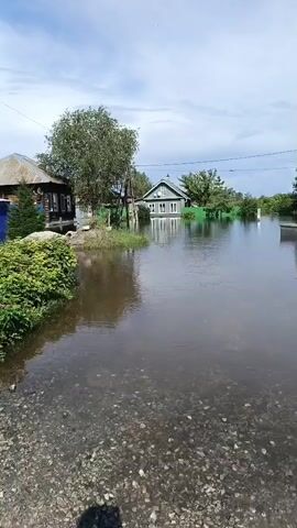 A natural disaster, likely a flood, has inundated a rural village. Water covers the ground, reaching the foundations of several houses. The sky is cloudy, suggesting recent or ongoing precipitation. The scene depicts the aftermath of a significant weather event, highlighting the vulnerability of rural communities to natural disasters.