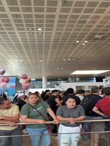 The video captures a busy indoor scene, likely an airport or a large public building, with a crowd of people. In the foreground, several individuals are visible, some looking towards the camera. In the mid-ground, more people are gathered, and there are displays, possibly departure boards, visible on the left. Several heart-shaped balloons, red and pink, are floating in the air, suggesting a celebration or a special occasion. The architecture of the building features a high ceiling with grid-like lighting fixtures and large glass windows. The overall atmosphere appears to be one of waiting or gathering.