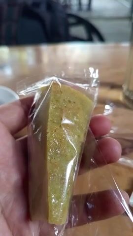 A hand holds a wedge-shaped piece of green cake in a clear plastic bag. The cake is textured with visible grains. The background is a blurred wooden table with a dark backpack and a white bowl. The bag is twisted at the top.
