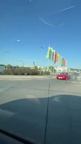 The video shows a first-person view from inside a car driving on a road under a clear blue sky. The camera is positioned facing forward through the windshield, with the dashboard and a side mirror visible. The car is moving along a paved road with multiple lanes. On the right side of the road, there are several large buildings with colorful banners and signs, likely commercial establishments or a shopping area. There are also streetlights and some greenery, including bushes and trees. In the side mirror, other cars can be seen. The overall scene depicts a sunny day in what appears to be a suburban or commercial district.