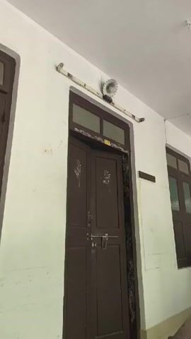 A video shows a dark brown double door with two panels on each side. Above the door, there is a light fixture attached to a white bar. To the right of the door, there is a window with brown panes. The walls are white. A ceiling fan is visible in the upper right corner of the frame.