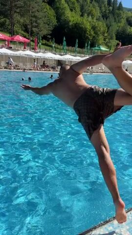 A person is captured mid-air performing a flip into a swimming pool. Water splashes around them as they descend. In the background, other people are swimming and relaxing by the pool. Lounge chairs and umbrellas are set up along the poolside, and a wooded hill with trees forms the backdrop.