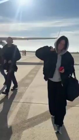 A woman wearing an oversized black jacket and black pants with a grey hoodie underneath is walking towards the camera, looking at a red phone in her hand. She is carrying a black bag over her shoulder. She stops walking and looks at the Ryanair airplane behind her. The video is shot on a sunny day on the tarmac. Another person walks past the woman in the opposite direction.