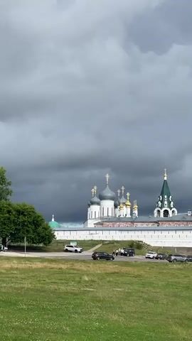 A static, vertical shot shows a large, white monastery with multiple domes and a green steeple. In front of the monastery, there is a white wall and a road with several cars parked. To the left of the monastery, a large green tree stands. A grassy field occupies the foreground, with a few small white flowers visible. In the middle ground, a body of water is visible to the right, with a boat docked. The sky is overcast with dark, ominous clouds.