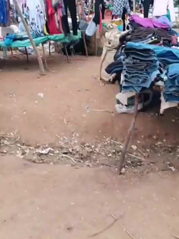The video shows an outdoor market stall with various items for sale, primarily clothing and shoes. In the foreground, several pairs of sneakers are laid out on a dark surface. Behind this, a woman sits in a green plastic chair, wearing a patterned skirt and a pink shirt. To her left, a woman in a green vest and black pants stands looking towards the stall. Another person, possibly a vendor, stands behind the seated woman, holding a pair of shoes. The stall itself displays a variety of colorful garments hanging on a makeshift rack. The ground is dusty and uneven, suggesting a rural or informal market setting. The sky is overcast, and there are some trees and structures in the background.