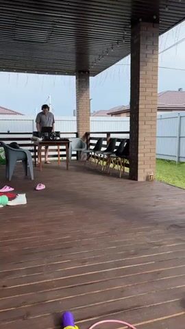 The video shows a backyard scene. In the foreground, there is a wooden deck with chairs and some scattered items. In the middle ground, there is a trampoline with two children jumping on it. To the right, there is a grass area with a blue inflatable pool. In the background, there are houses, fences, and two cars parked on a paved area. A person is visible on the deck, standing near a laptop. The weather appears to be overcast.
