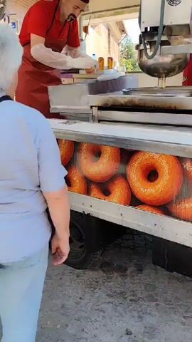 The video shows a donut food truck. A person wearing a red shirt and apron, along with white gloves, is preparing donuts behind the counter. The side of the truck is decorated with an image of numerous donuts. Another person, wearing a light blue shirt, stands in front of the truck, seemingly interacting with the person making donuts. The scene appears to be outdoors, with a gray concrete surface on the ground.